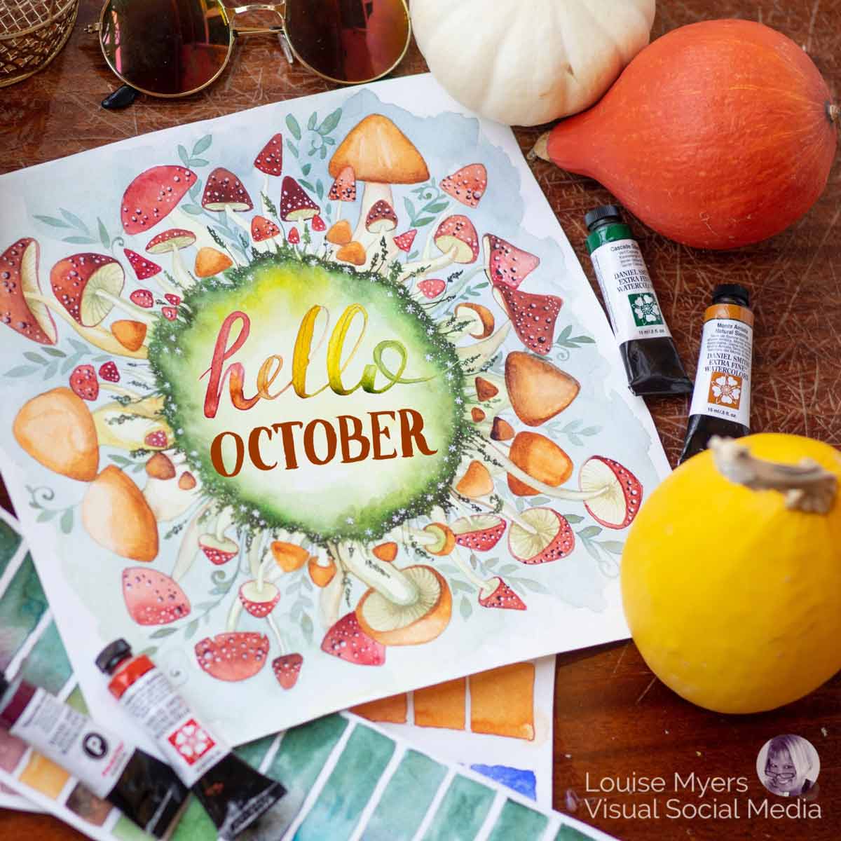 watercolor art of mushrooms surrounding words "hello october" with paints and gourds on wood tabletop.